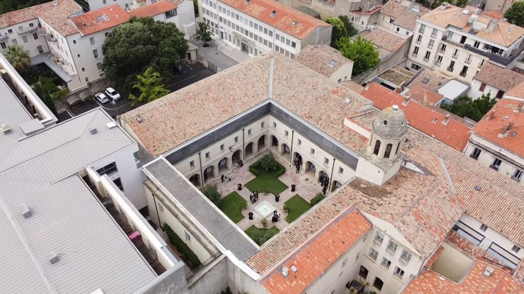 Vue aérienne du cloître historique de la Faculté de Droit de Montpellier, révélant une cour intérieure avec fontaine centrale et espaces verts, encadrée d’arcades en pierre et de bâtiments traditionnels aux toits de tuiles rouges