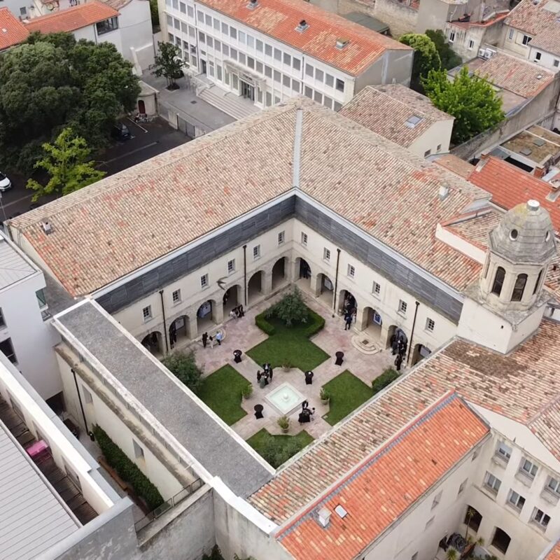 Aerial view of the historic cloister at Montpellier Law School, revealing an inner courtyard with a central fountain and green spaces, framed by stone arcades and traditional buildings with red-tiled roofs