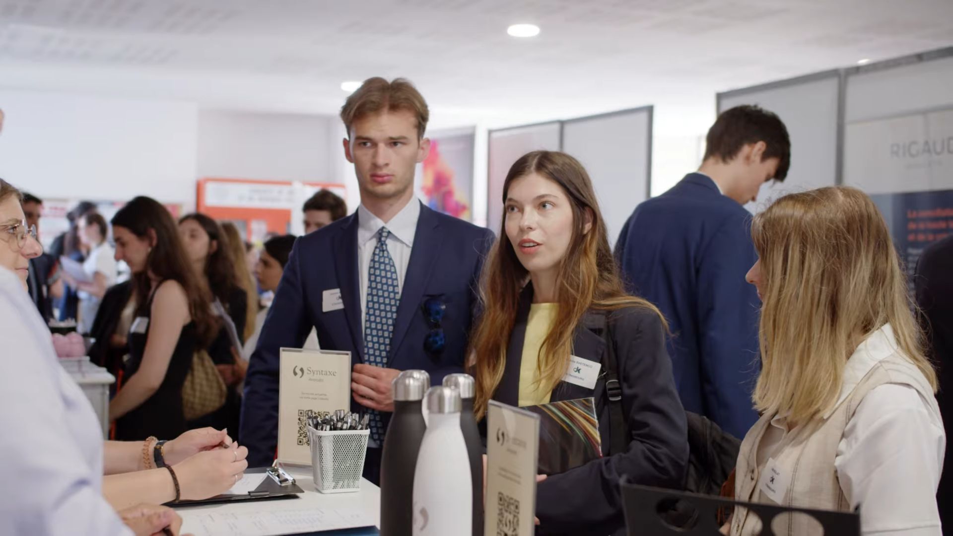Video screenshot from the DJCE Job Fair 2025, showing a group of law students gathered in front of the Syntaxe booth, with branded firm goodies on display, as they engage in a lively conversation with the recruiters in the bustling recruitment fair