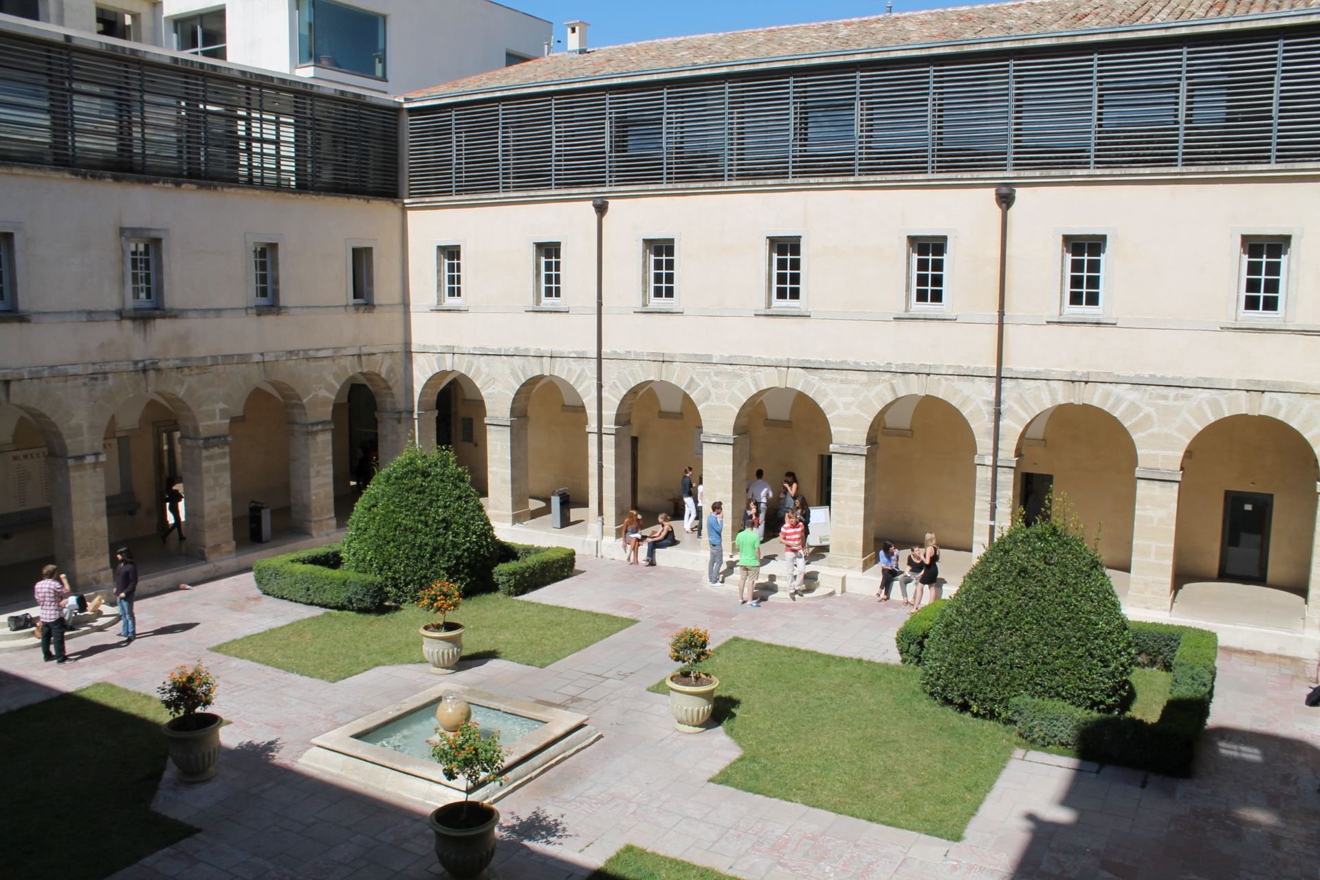 Vue du cloître historique de la Faculté de Droit de Montpellier, focalisée sur l’angle sud-ouest, illuminé par le soleil, avec sa fontaine centrale, ses haies géométriques et ses arcades en pierre